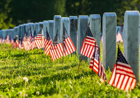 Rows of white headstones with small American flags in a sunlit cemetery