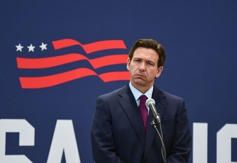 A public figure standing at a microphone with a serious expression against a backdrop featuring the USA flag
