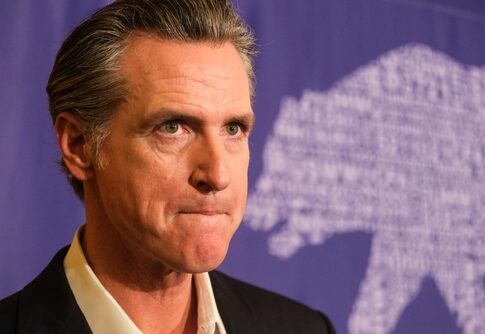 A serious-looking political figure at a press event with a bear graphic in the background